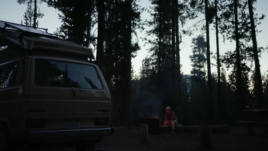 Person sits next to small campfire and camper van in forest at night