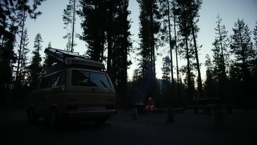 Person sits on log near campfire in forest clearing near parked van