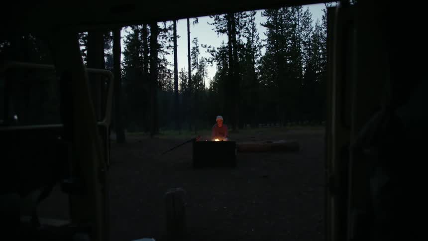 A camper sits on a log next to a bonfire as his vehicle is parked nearby with its door wide open