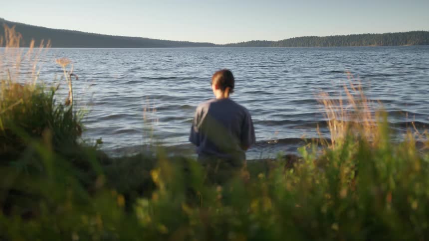 A young woman sits on the shore of a lake during the day