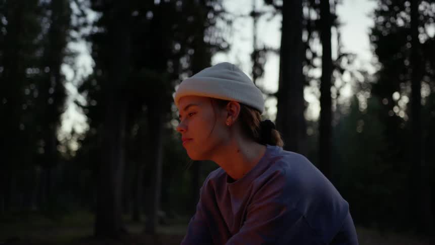 Woman with a white beanie and a sweatshirt sits by a campfire in the forest at dusk