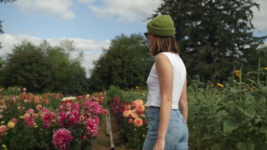 A woman in a beanie and sunglasses walks past rows of flowers in a field during the day