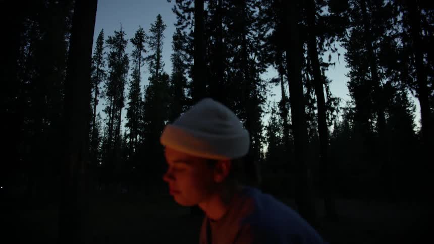 A woman sits in front of a fire in a woodland in the evening