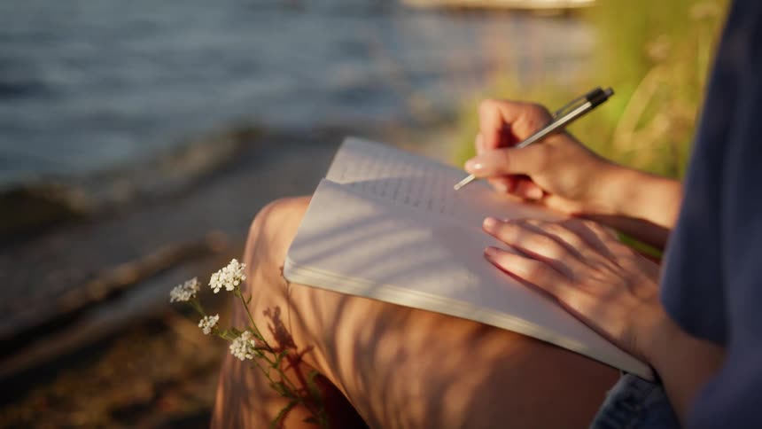 A person sits by the water edge writing in a notebook