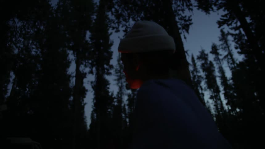 Woman with a white beanie stands next to the campfire in the woods at dusk