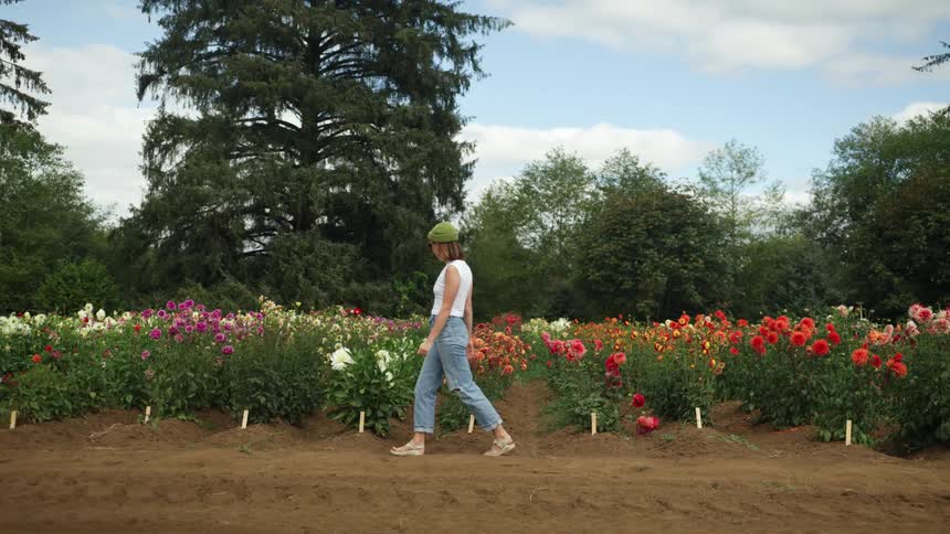 Woman walks past rows of flowers bordered by trees under cloudy sky