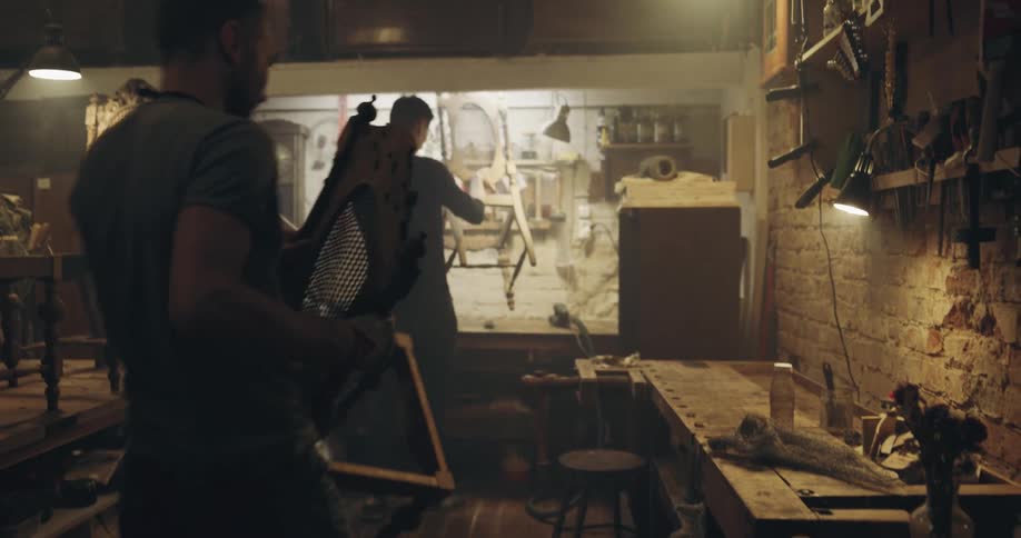 Two men carrying wooden chairs in a workshop