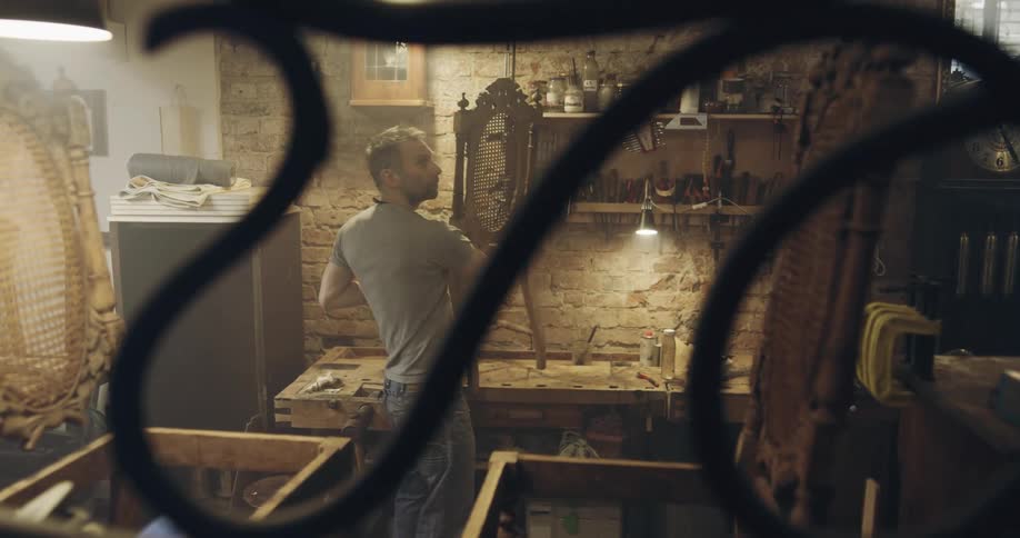A man and teenager carry old chairs into a dim workshop