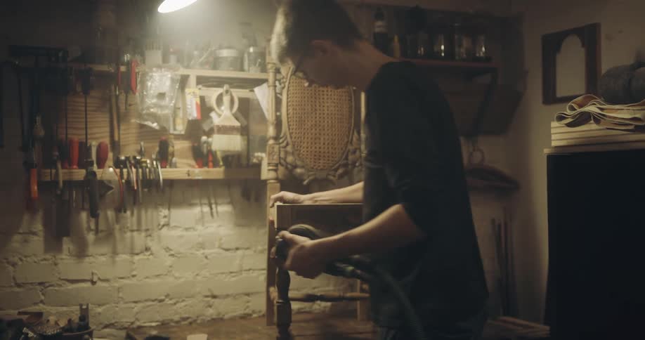 A man in a workshop sanding a wooden chair with tools in the background