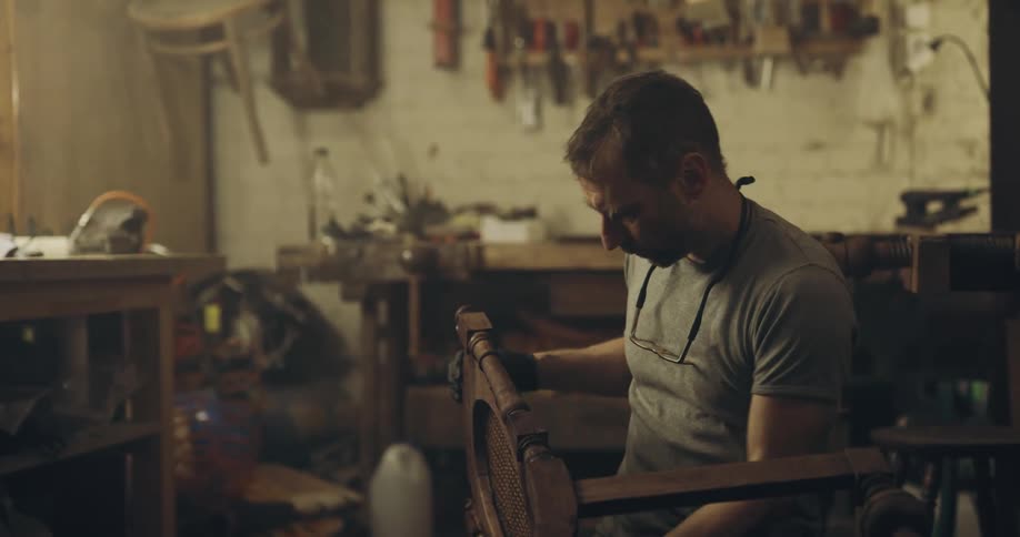 A man sits in a dim workshop painting a chair