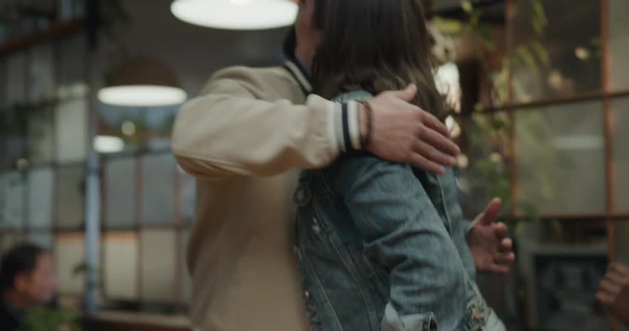 A woman sits at a table at a restaurant talking to a man and then another woman walks up with a group and covers her eyes from behind and they all say hello and hug
