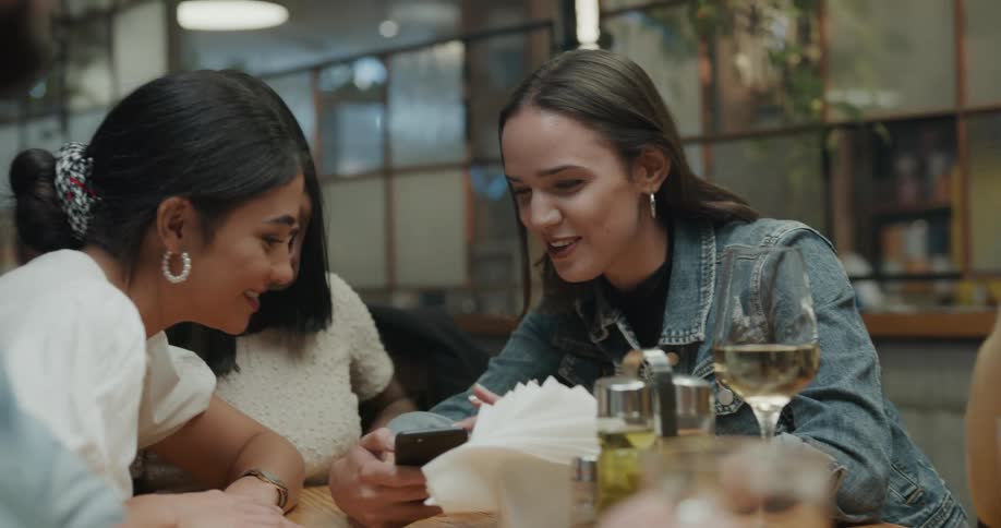 3 female friends sit at a table in a restaurant with wine and look at something on a phone together