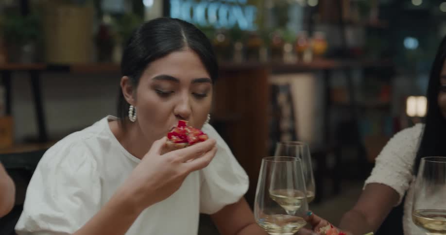 A group of friends sitting at a bar with glasses of wine each take a piece of bruschetta and eat it