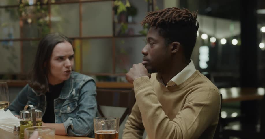 Friends sit around table in bar with glasses of beer laughing and talking