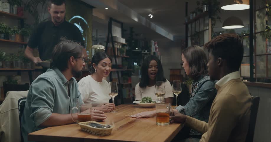 A group of people sitting together in a restaurant chat while a server places food on the table before them