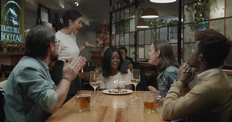 A waitress places a slice of cake in front of a woman sitting with her friends at a table in restaurant and leads the womans friends in singing to her