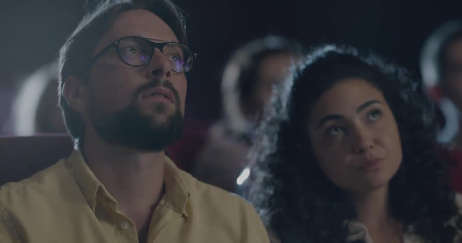 Woman with curly hair and man with a beard and glasses eat popcorn and watch a movie in the movie theater