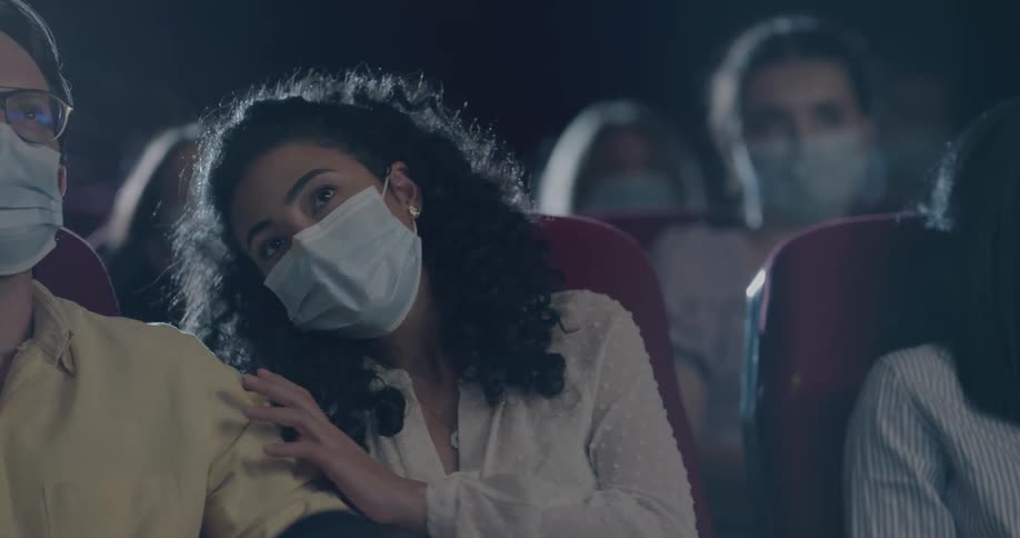 Woman taps on man and gestures to the screen while watching a movie in a theater with moviegoers wearing masks