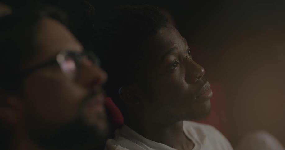 White man with a beard and glasses and black man with a white t shirt watch a movie while eating popcorn in the movie theater