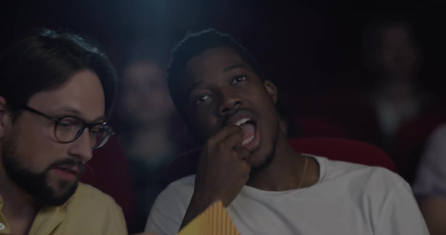 Black man and white man eat popcorn and watch a movie in the movie theater