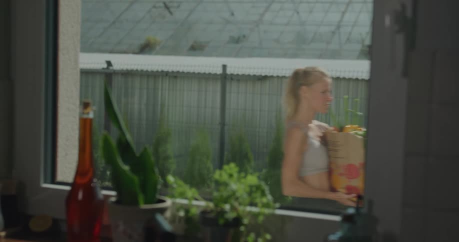 A woman walks past a window a home carrying a bag of groceries