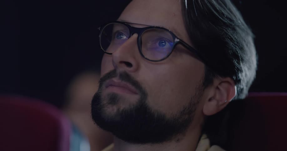 A man wearing glasses looks up at the screen while sitting in a cinema theater
