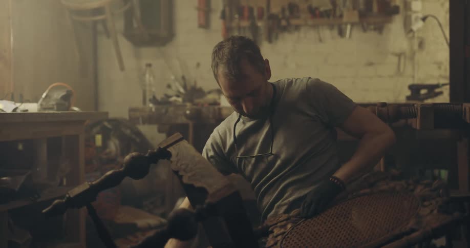 Man wipes parts of wicker and wood chair crouching on floor of workshop near brush