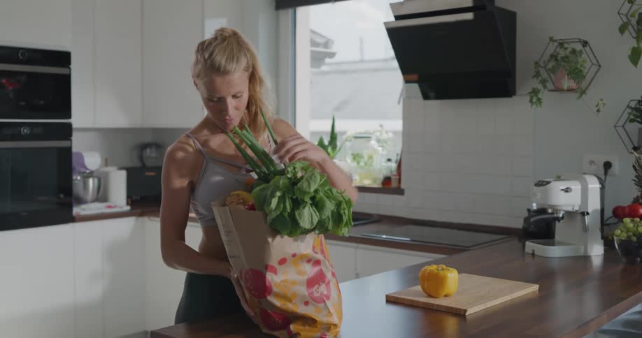 A woman carries a bag of groceries into her kitchen and begins to unpack its contents onto a wooden cutting board on the counter