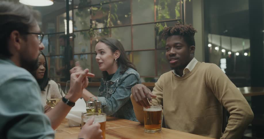 Friends sit around table with alcoholic drinks laughing and talking