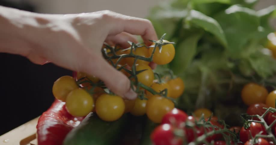 Person picks up red cherry tomatoes still on the vine from among other produce on chopping board