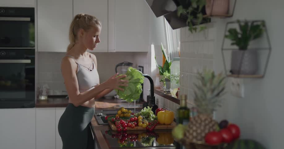 A woman in athletic wear washes fruits and vegetables under a kitchen faucet
