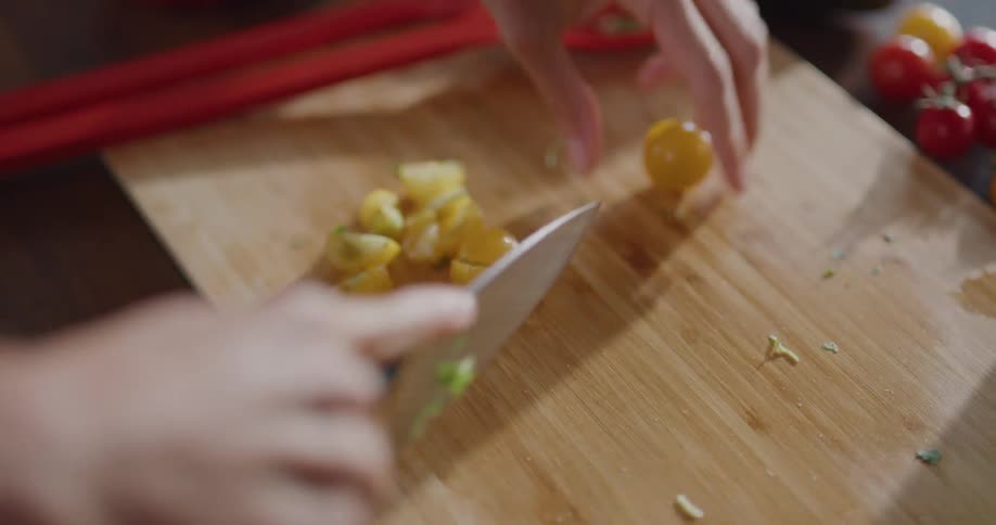 A person cuts small cherry tomatoes and adds them to a salad bowl