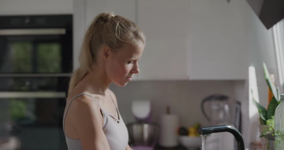 Blonde woman wearing workout gear washes produce at kitchen sink
