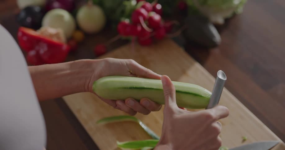 A person stands in the kitchen and peels the skin off of a cucumber