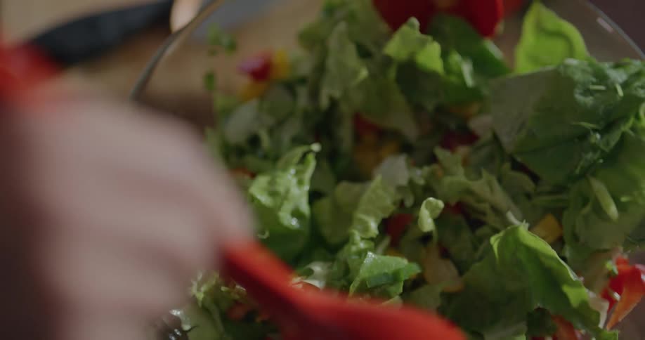 A person uses a pair of large spoons to toss a salad in a glass bowl