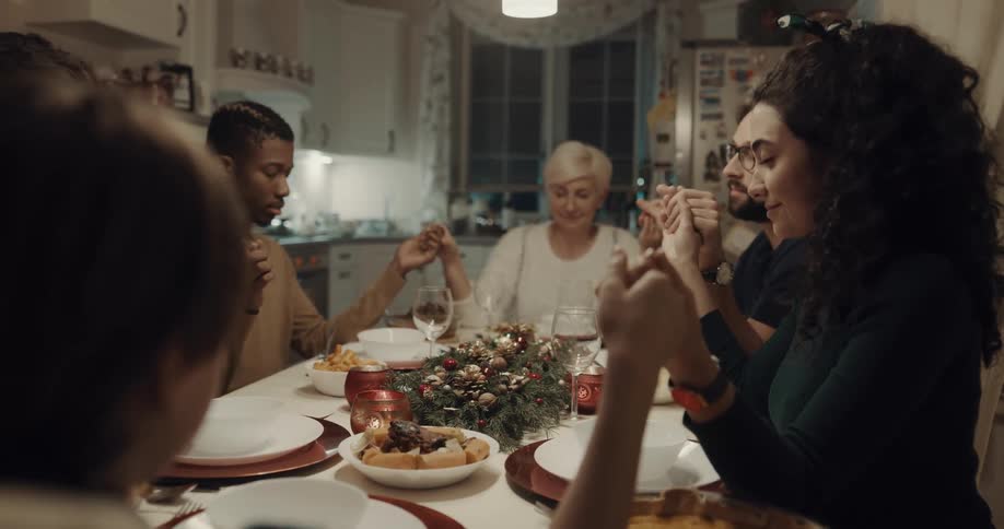 A group of friends are gathered around the dining room table that is decorated for christmas and join hands to begin praying before sharing a meal together