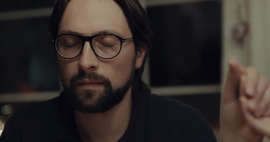 A man sits with his head bowed in prayer while holding hands with another person as grace is said at a dining table before opening his eyes and talking once it has finished