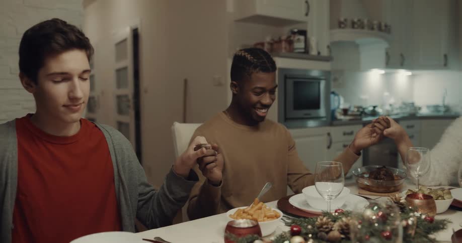 A group of people hold hands and bow their heads as grace is said at a dining table in a home kitchen