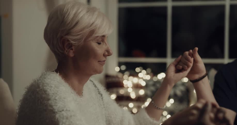 A white haired woman holds the hands of two other people while closing her eyes in prayer