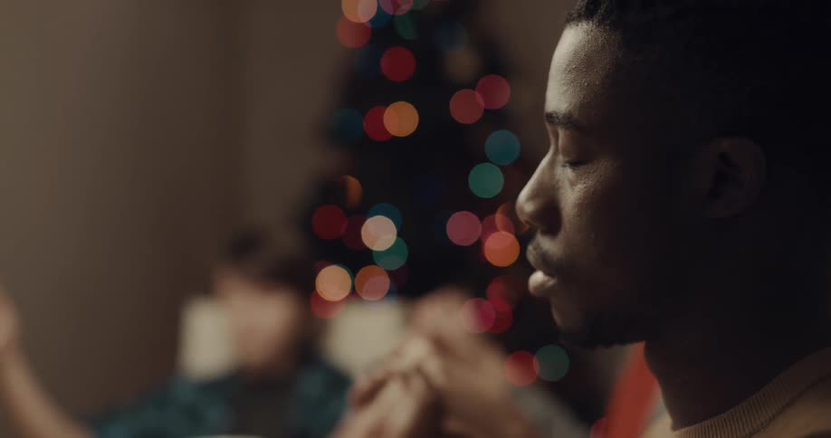 Family and friends clasp hands to pray at dinner table with christmas tree in background