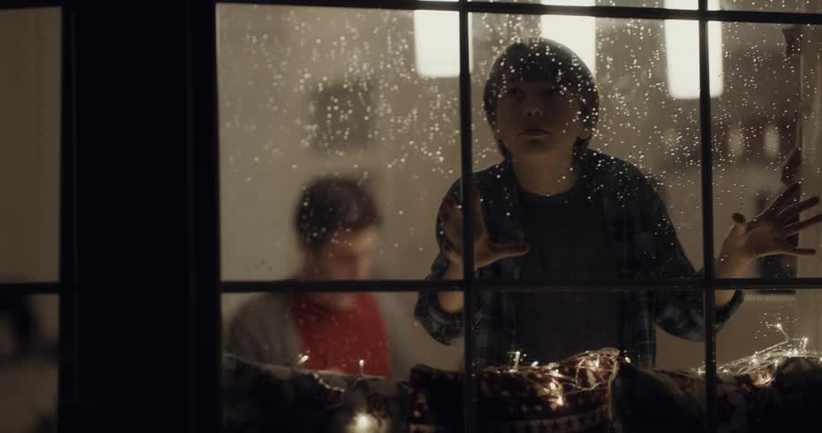 A boy looking out of the window with droplets with people sitting at a table in the background