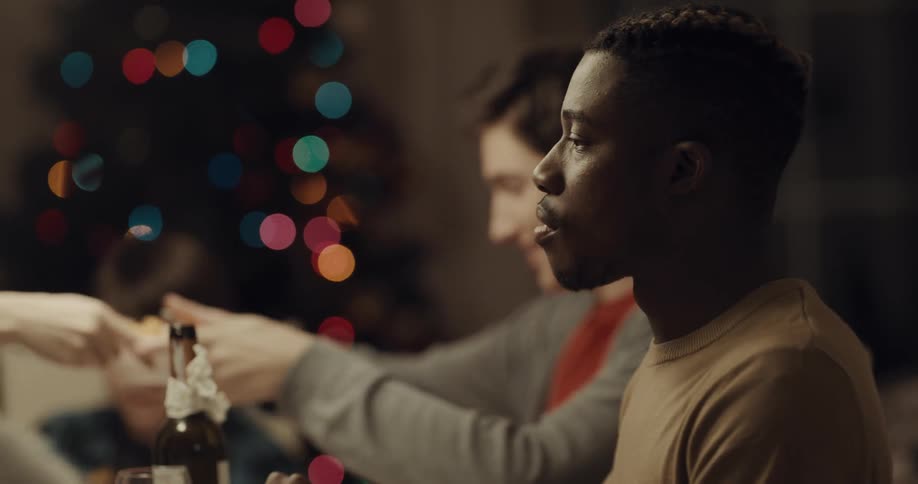 A group of people sit around a home dinner table eating and passing food to each other with a christmas tree in the background