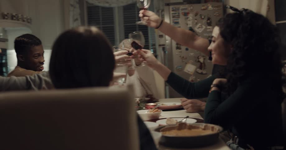 A group of people raise their wineglasses for a toast as they cheerfully converse at a dining table in a home kitchen