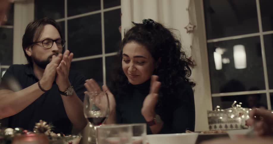 As a family sits at a dinner table and eats a man puts a spoon of food in to his girlfriends mouth and they laugh