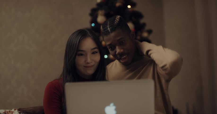 A man and woman stand in front of a christmas tree and look at a laptop and talk and laugh