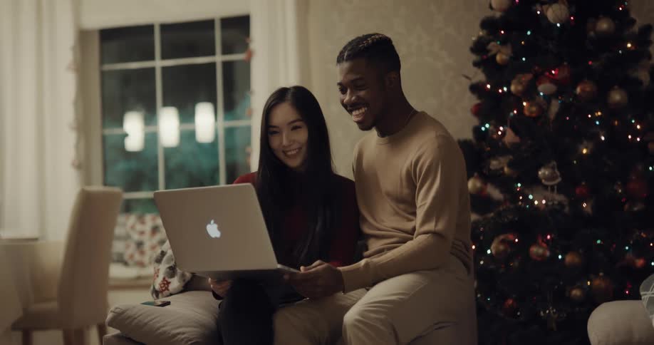 A man and woman smile and laugh while looking at the screen of a laptop as they sit on a sofa in front of a christmas tree in the living room of a house at night