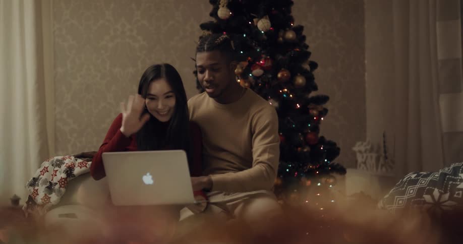 A man and woman sit together having a video call on a laptop with a christmas tree behind them