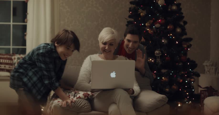 Woman with short grey hair sitting in an armchair next to the christmas tree makes a video call on a laptop and asks kids to come in the living room at home