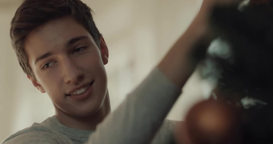 A young man places decorations on a christmas tree while talking to someone out of frame