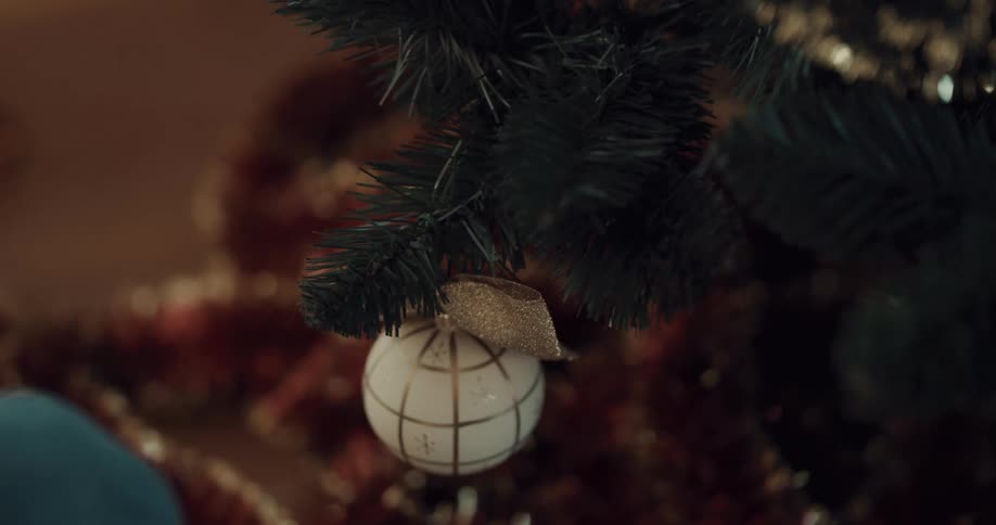 A white young person wearing a red plaid outfit is hanging christmas ornaments on a green christmas tree